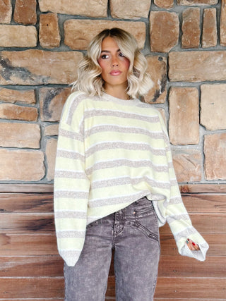 A woman with blonde wavy hair wearing a yellow and taupe striped knit sweater and brown jeans, standing in front of a rustic stone and wood wall.