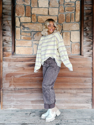 A woman with blonde wavy hair wearing a yellow and taupe striped knit sweater and brown jeans, standing in front of a rustic stone and wood wall.