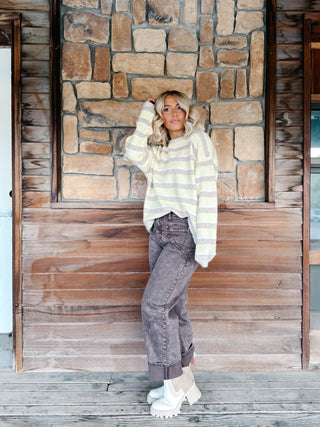 A woman stands on a wooden porch in front of a rustic stone and wood wall, wearing a yellow and mocha striped knit sweater, faded brown jeans, and white platform boots.