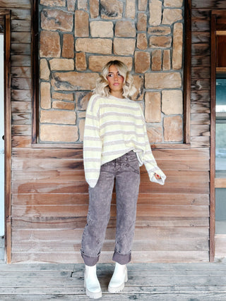 A woman stands on a wooden porch in front of a rustic stone and wood wall, wearing a yellow and mocha striped knit sweater, faded brown jeans, and white platform boots.