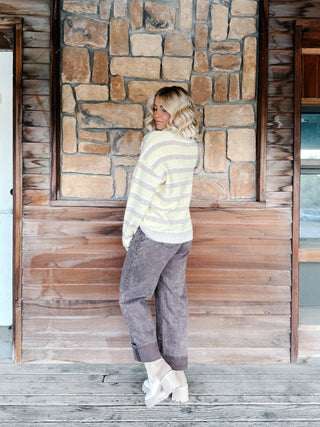 A woman stands on a wooden porch in front of a rustic stone and wood wall, wearing a yellow and mocha striped knit sweater, faded brown jeans, and white platform boots.