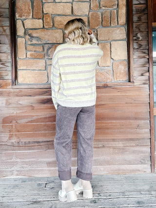A woman stands on a wooden porch in front of a rustic stone and wood wall, wearing a yellow and mocha striped knit sweater, faded brown jeans, and white platform boots.