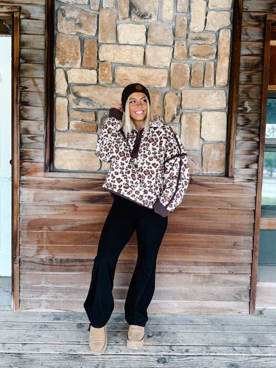 A woman wearing a brown and beige leopard print fleece pullover hoodie with dark brown trim, paired with black pants and a dark brown beanie, standing in front of a rustic stone wall.