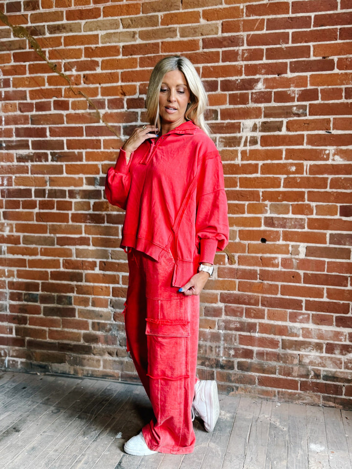 A woman wearing a matching red lounge set consisting of a hooded sweatshirt and cargo pants, standing against a brick wall.