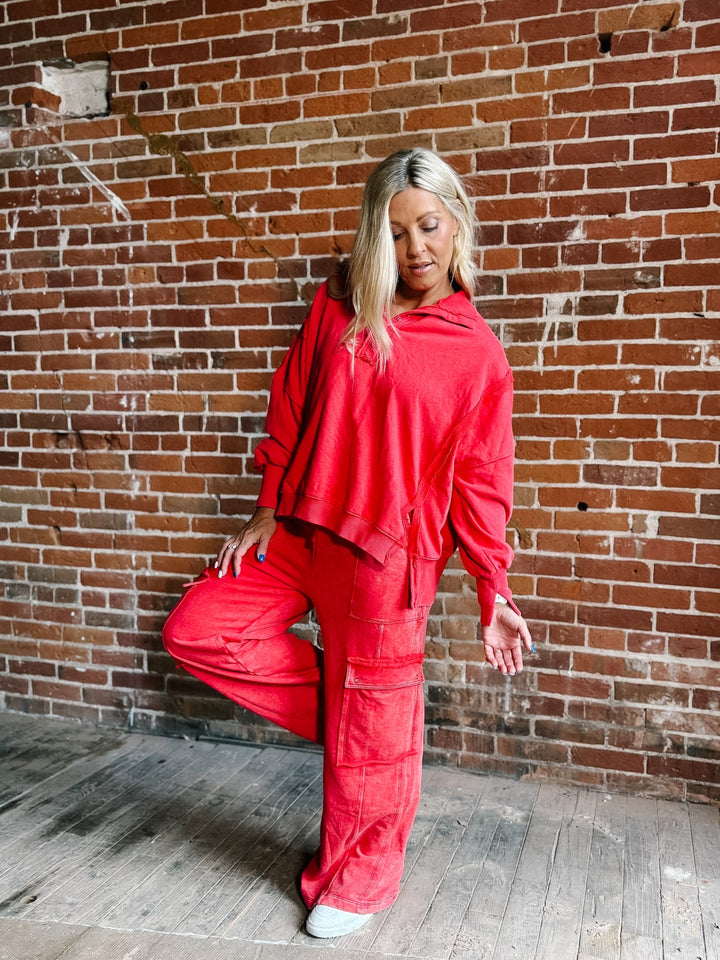 A woman posing in a matching bright red two-piece lounge set consisting of a cropped long-sleeve top and cargo-style pants, set against a rustic red brick wall background.