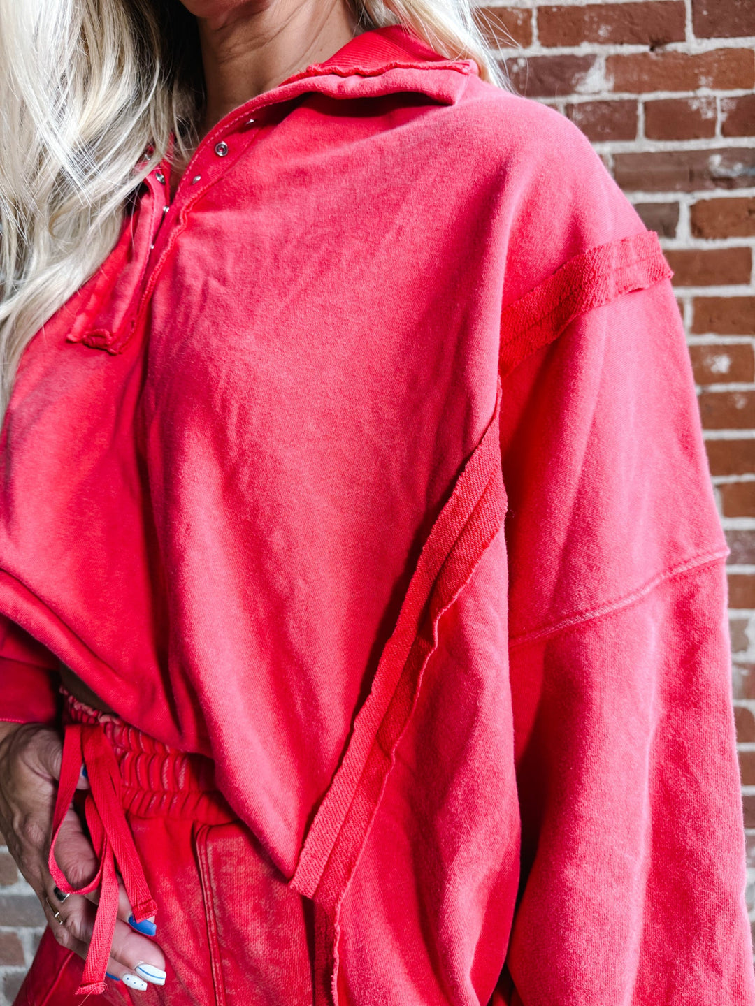 A close-up of a person wearing an oversized red hooded sweatshirt or lounge set in front of a brick wall. The garment has a soft, slightly faded texture and includes a drawstring waist.
