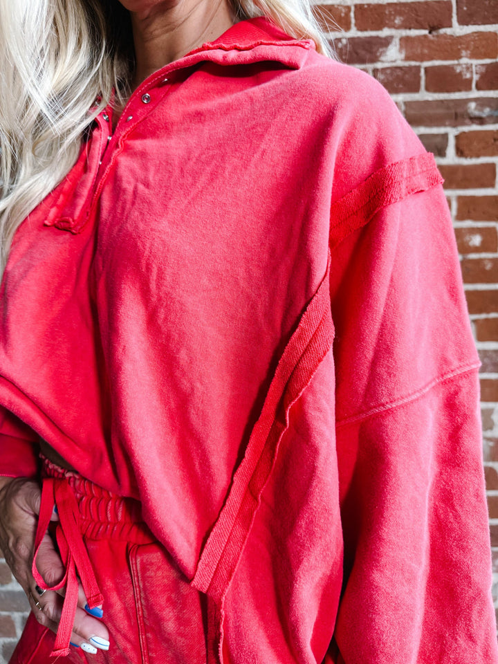 A close-up of a person wearing an oversized red hooded sweatshirt or lounge set in front of a brick wall. The garment has a soft, slightly faded texture and includes a drawstring waist.