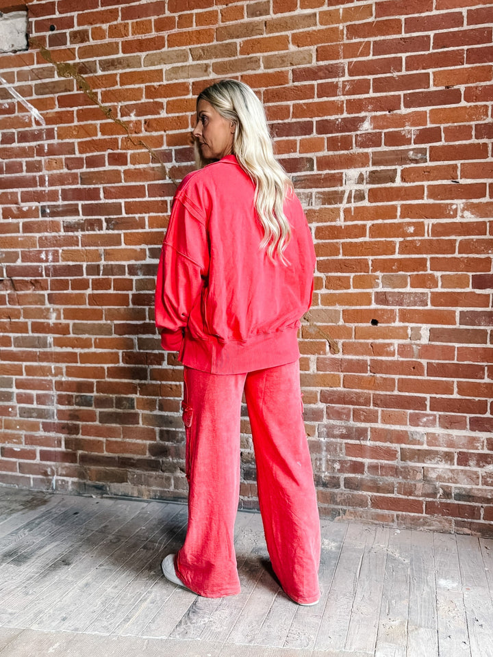 A woman with blonde hair stands with her back to the camera, wearing a matching bright coral-red sweatshirt and wide-leg pants set, posed in front of a red brick wall.