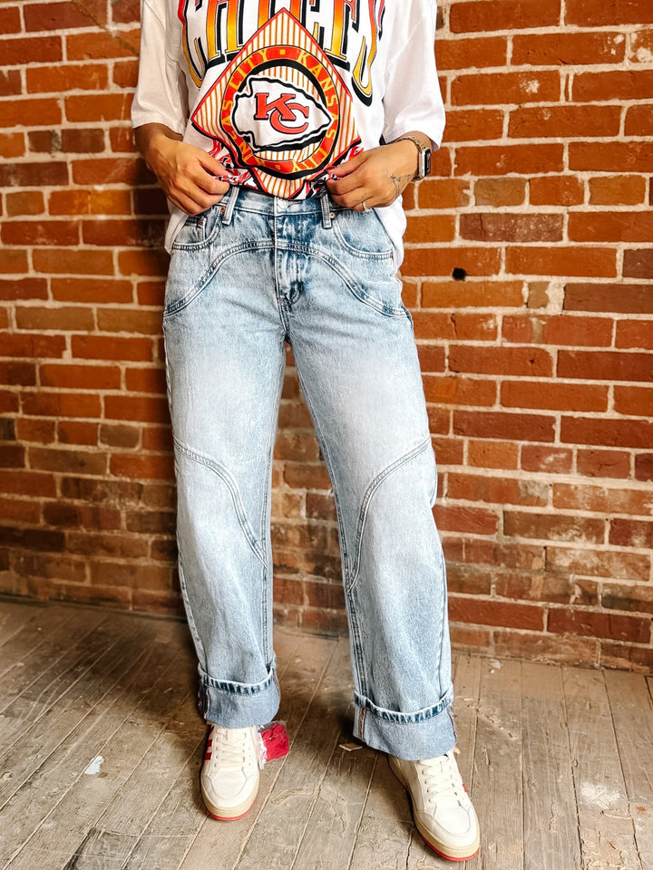 A person wearing light-wash blue wide-leg denim jeans, a white Kansas City Chiefs graphic t-shirt, and white sneakers, standing in front of a red brick wall.