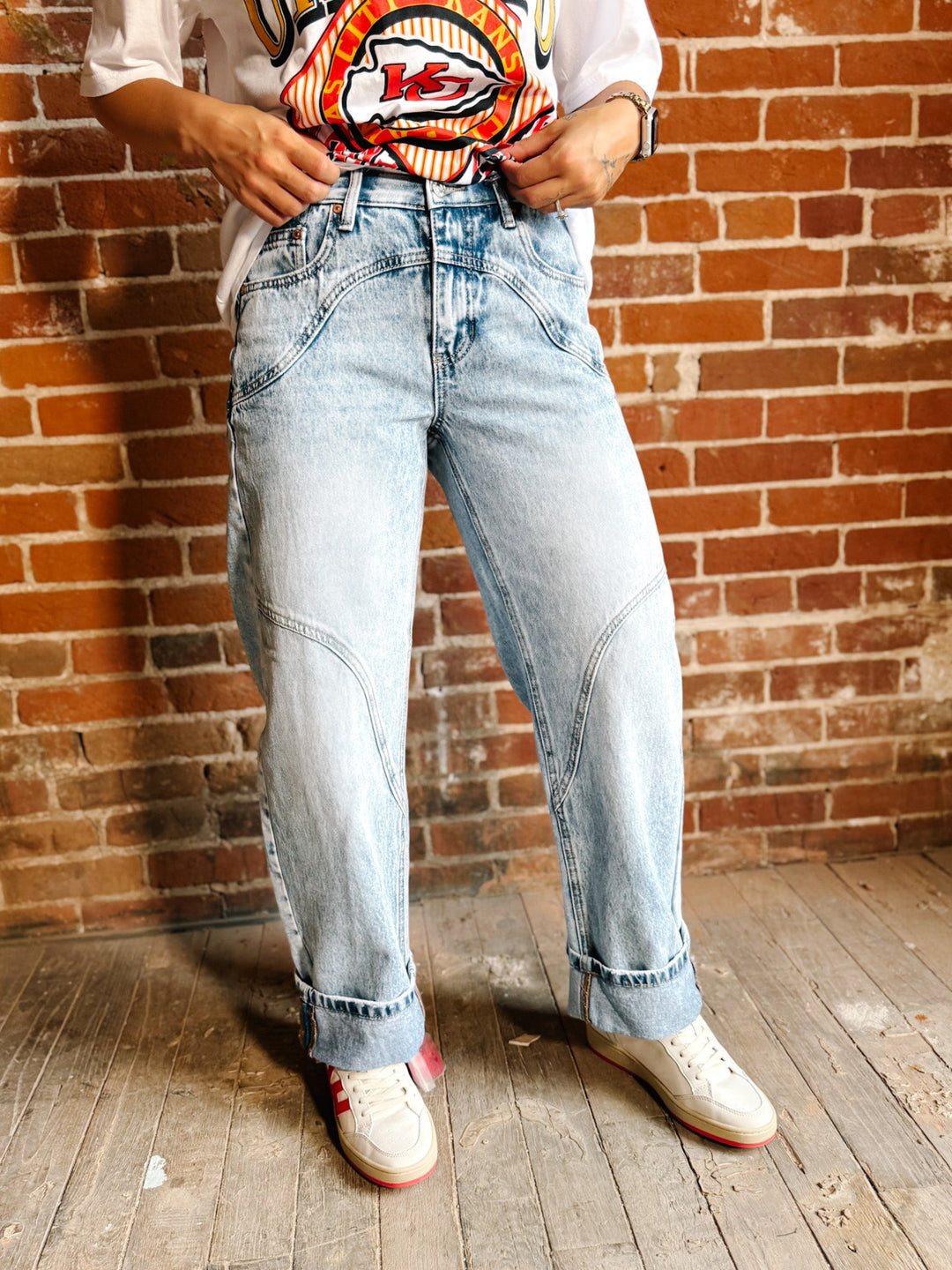 A person wearing light-wash blue denim jeans with a vintage graphic t-shirt and white sneakers, standing in front of a red brick wall.