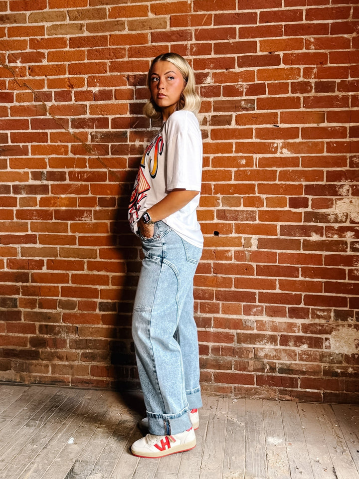 A woman stands against a red brick wall wearing a white graphic t-shirt, light-wash wide-leg jeans, and white sneakers with red accents.