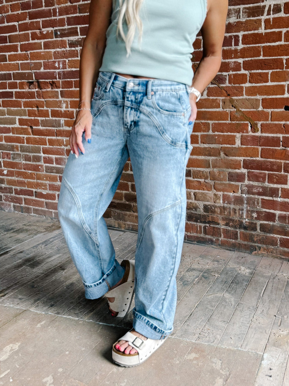 A person wearing light-wash wide-leg denim jeans, a pale mint green tank top, and white slide sandals, standing against a red-brown brick wall.