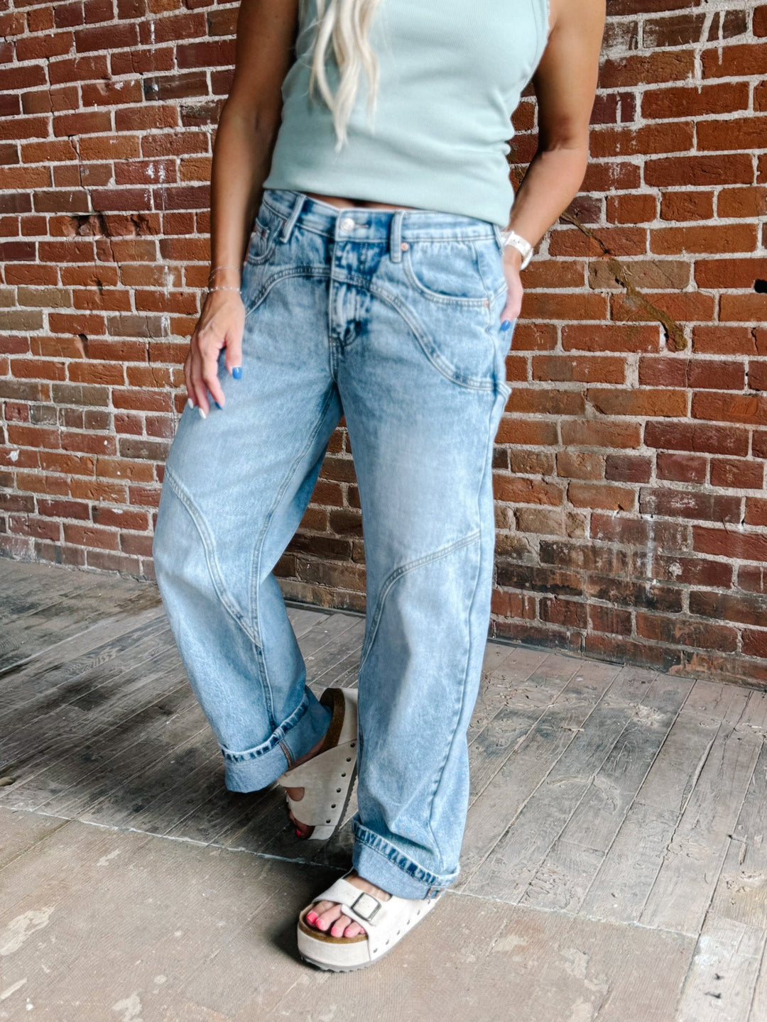 A person wearing light-wash wide-leg denim jeans, a pale mint green tank top, and white slide sandals, standing against a red-brown brick wall.