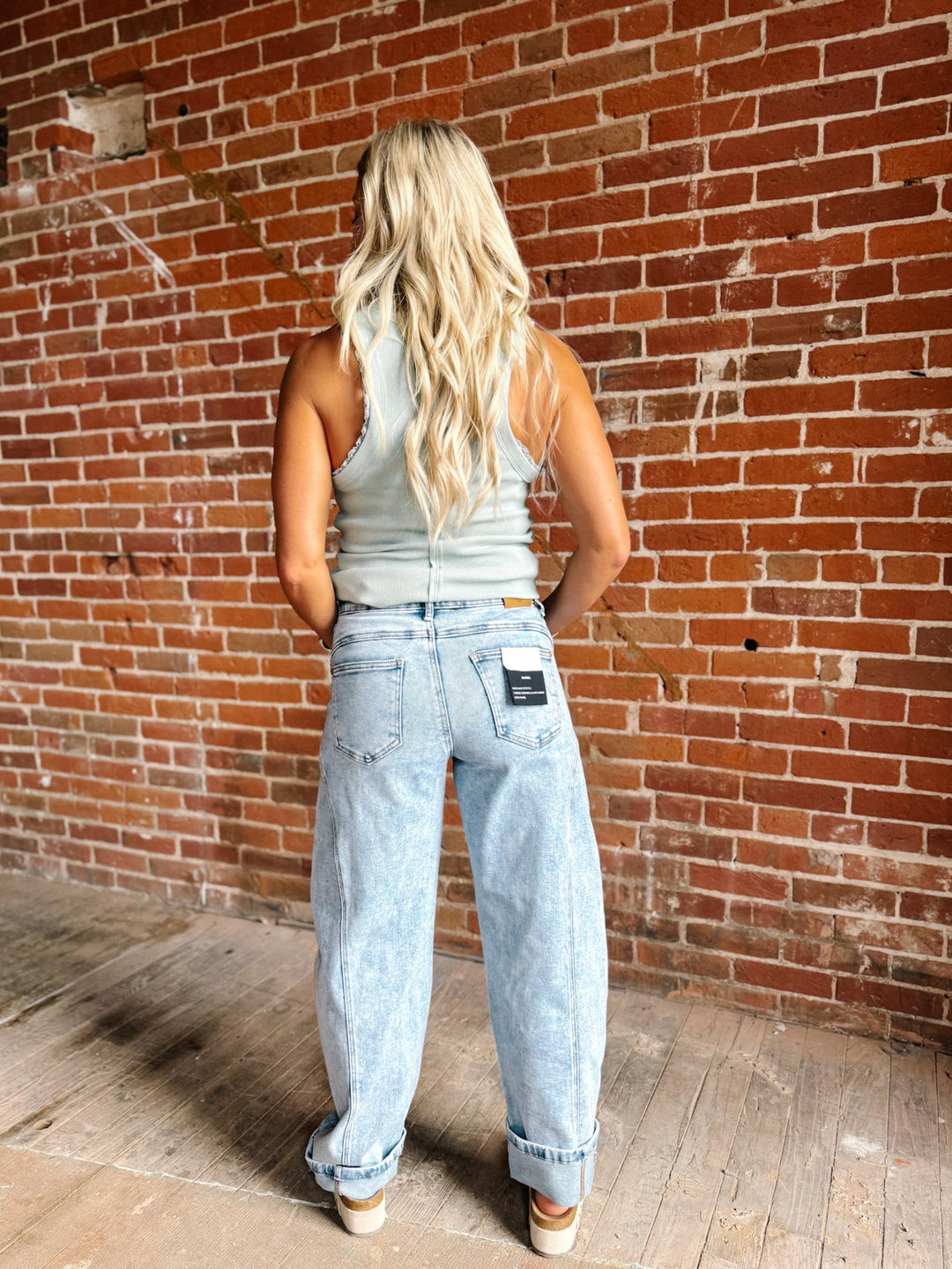 A woman with long blonde hair is shown from behind wearing light-wash, wide-leg denim jeans and a light gray tank top, standing in front of a red brick wall.