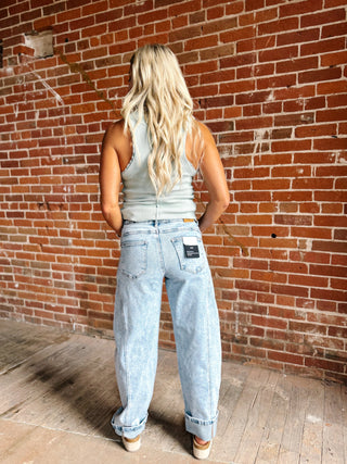 A woman with long blonde hair is shown from behind wearing light-wash, wide-leg denim jeans and a light gray tank top, standing in front of a red brick wall.