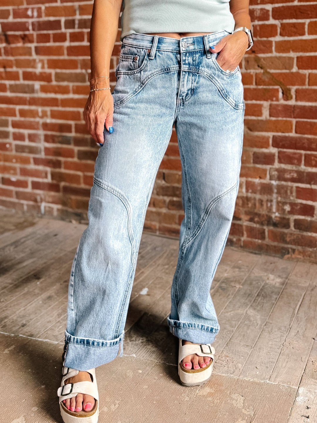 A person wearing light-wash wide-leg denim jeans with cuffed hems, standing against a red brick wall and wooden floor.