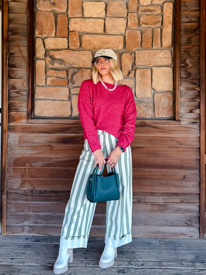A woman stands in front of a rustic wooden and stone wall, wearing a bright pink knit sweater, green and white vertically striped wide-leg pants, white platform boots, a beige baseball cap, and carrying a teal handbag.