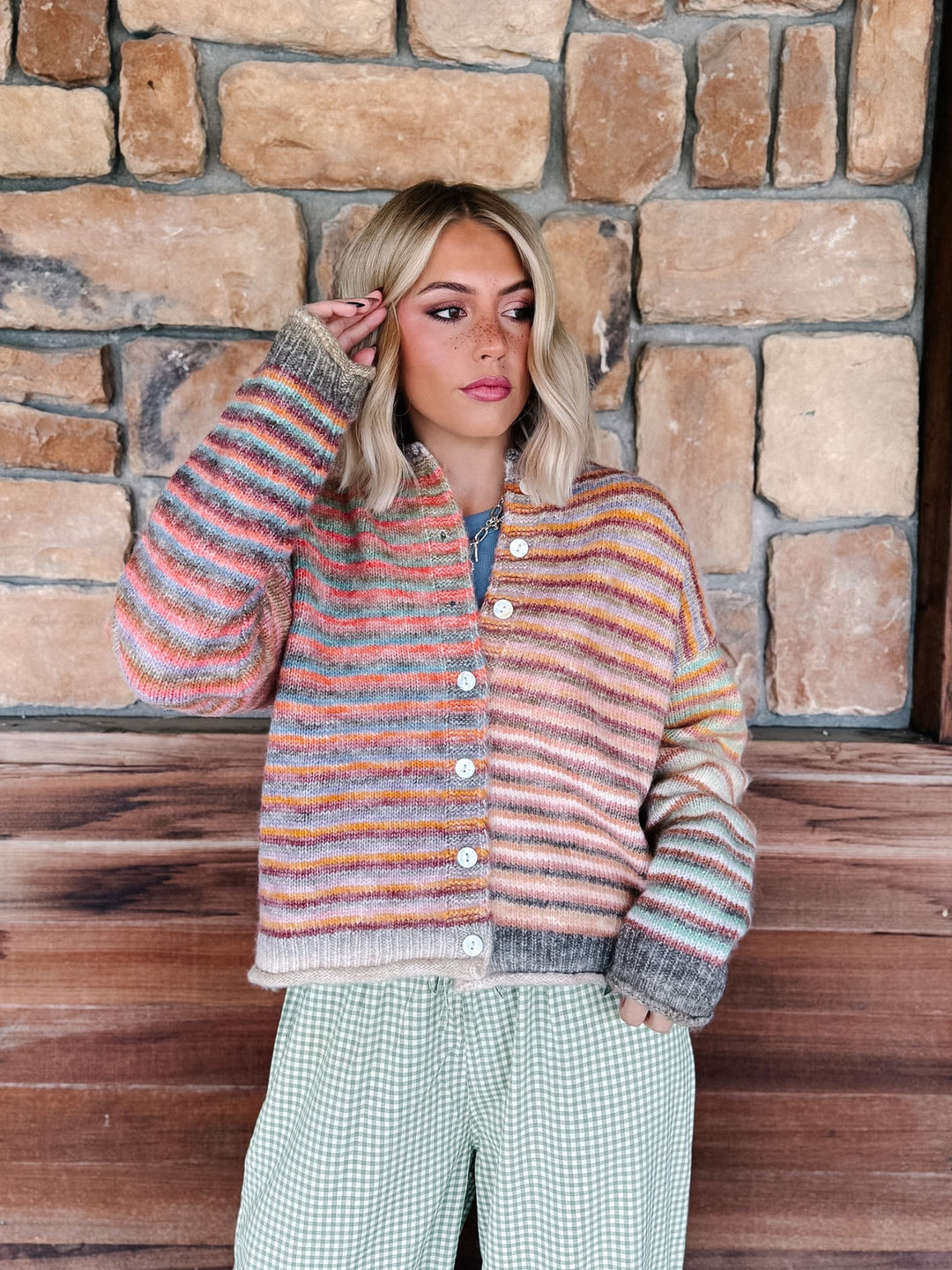 A woman stands in front of a rustic stone wall wearing a fuzzy striped cardigan with pink, orange, blue, and gray tones, paired with green-and-white gingham pants.