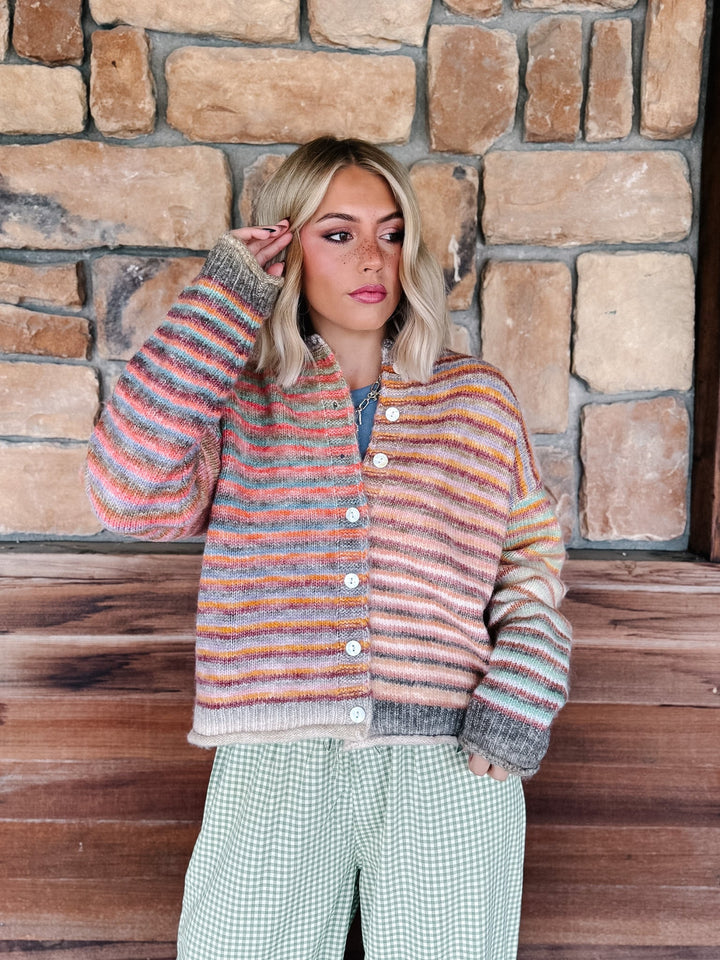 A woman stands in front of a rustic stone wall wearing a fuzzy striped cardigan with pink, orange, blue, and gray tones, paired with green-and-white gingham pants.