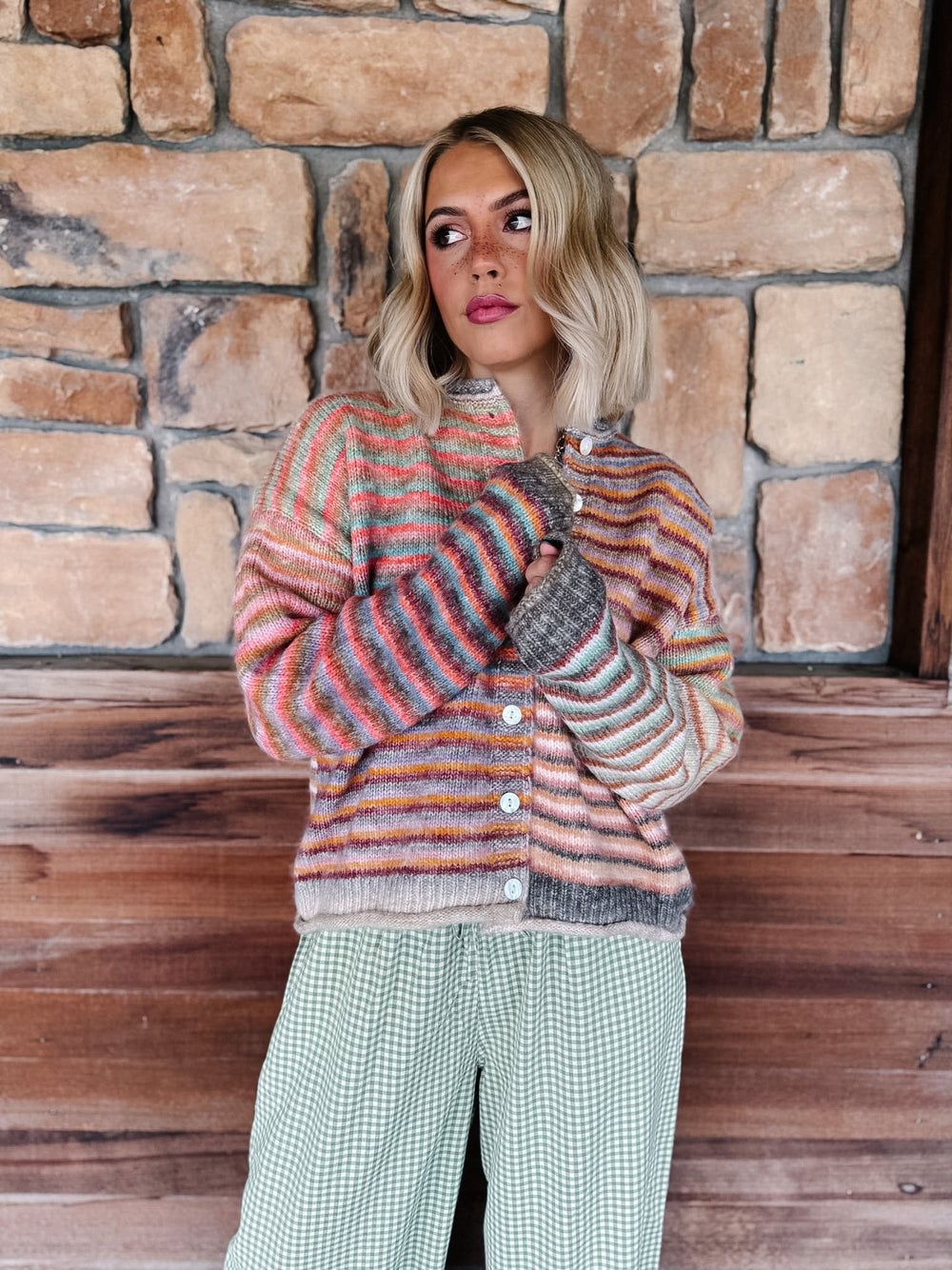 A woman wearing a striped knit cardigan with a half-light, half-dark color-block design, paired with green and white checkered pants, standing in front of a rustic stone wall.