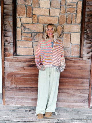 A woman stands on a rustic wooden porch in front of a stone wall, wearing a striped knit cardigan in peach, orange, and blue-gray tones with wide-leg sage green pants and tan boots.
