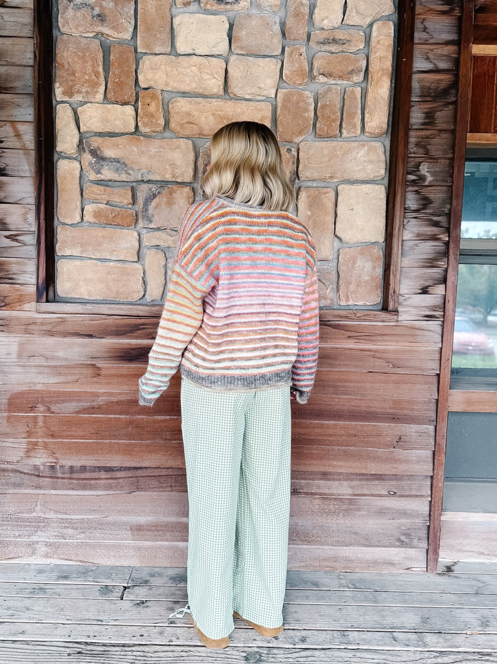 A woman stands with her back to the camera wearing a striped knit sweater and wide-leg green pants, posed in front of a rustic stone and wood wall.
