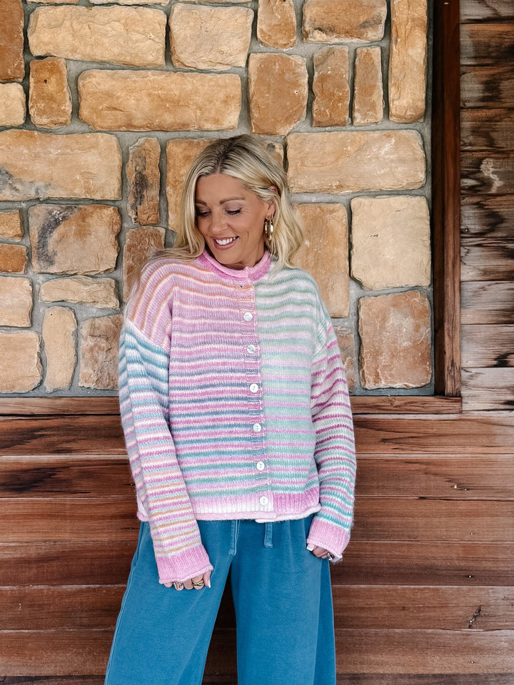 A woman wearing a pastel striped knit cardigan sweater and blue jeans, standing in front of a rustic stone and wood wall.