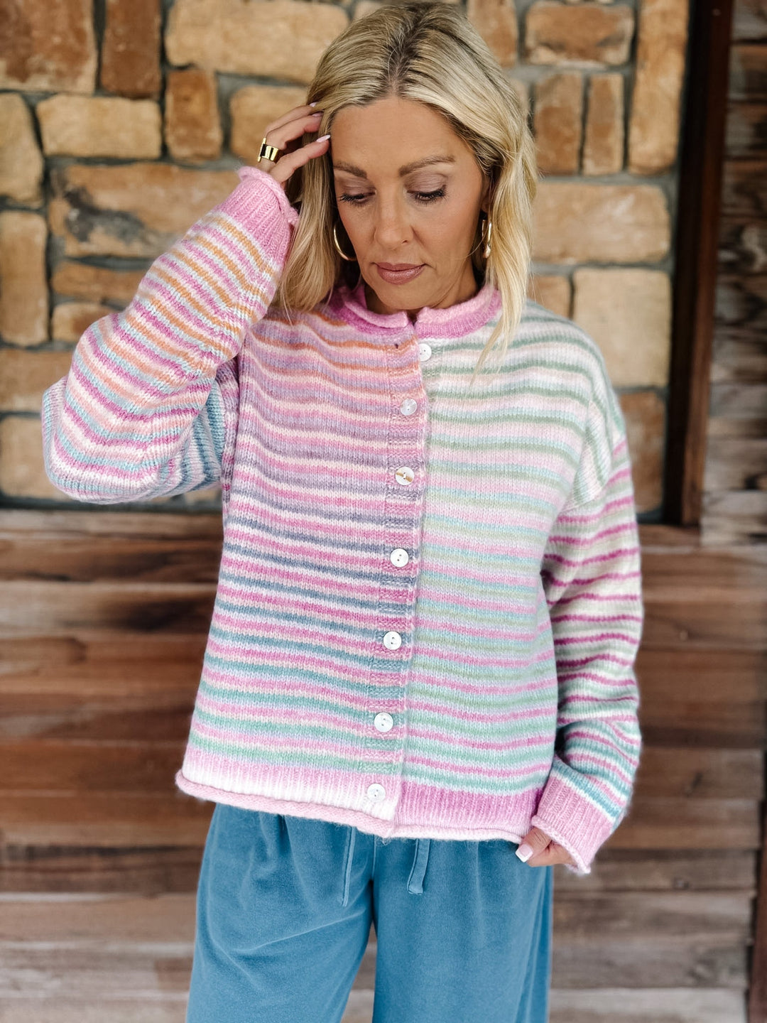 A woman wearing a pastel striped knit cardigan sweater with pink, blue, and yellow stripes, paired with light blue pants, standing in front of a rustic stone and wood wall.