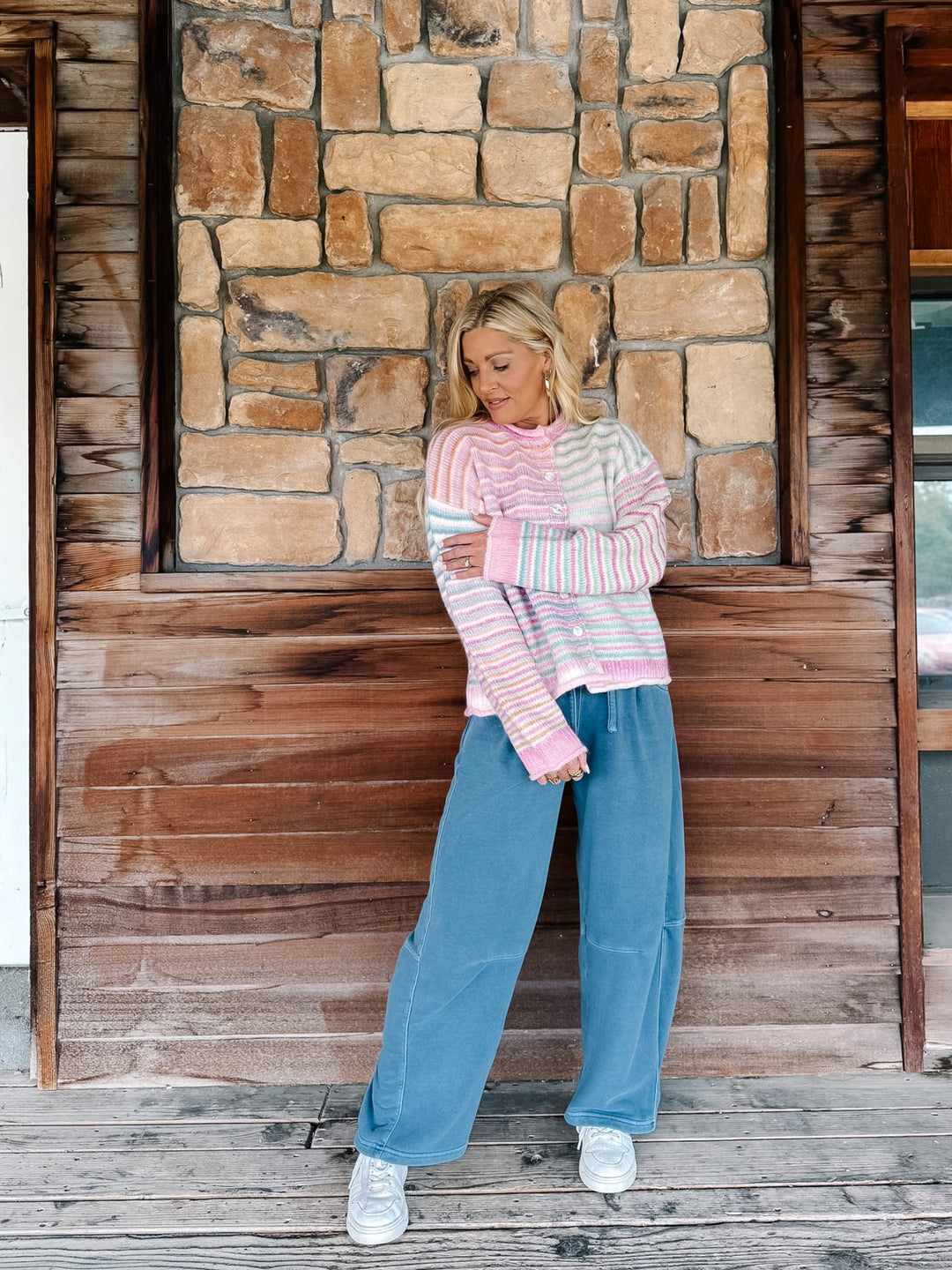 A woman stands on a rustic wooden porch in front of a stone wall, wearing a pink and white striped long-sleeve crop top, wide-leg blue jeans, and white sneakers.