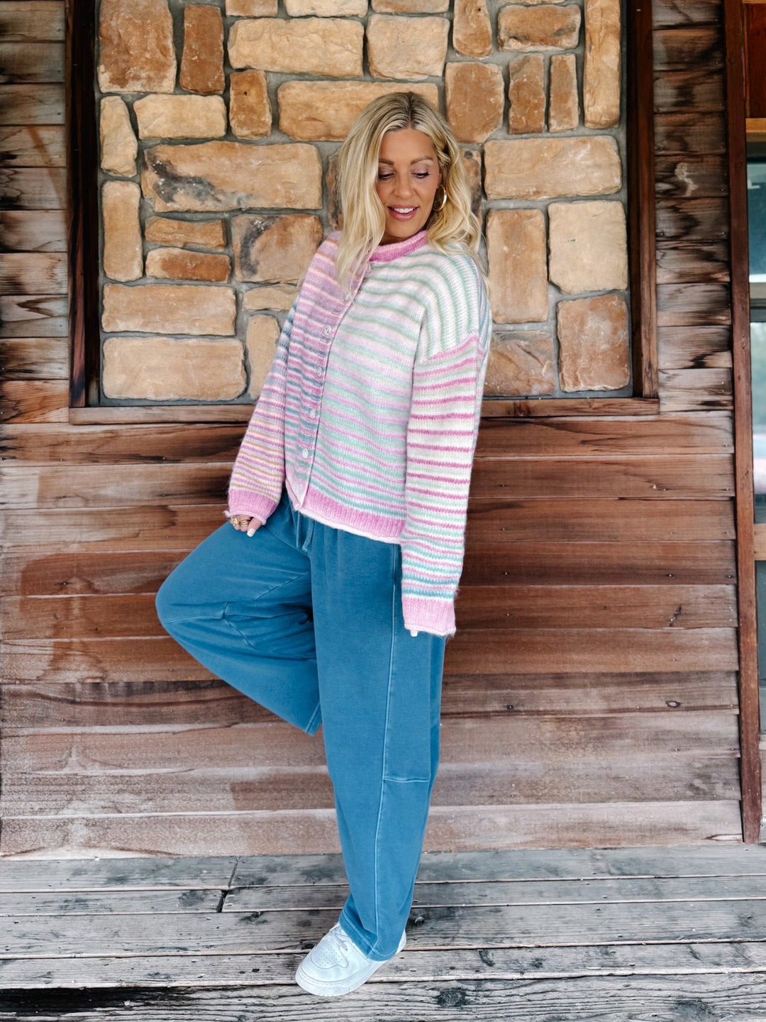 A woman posing on a rustic wooden porch in front of a stone wall, wearing a pastel striped cardigan with pink and white stripes, blue jeans, and white sneakers.