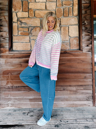 A woman posing on a rustic wooden porch in front of a stone wall, wearing a pastel striped cardigan with pink and white stripes, blue jeans, and white sneakers.