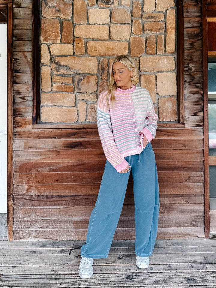 A woman stands on a wooden porch in front of a stone wall, wearing a pastel striped cardigan with pink, white, and light blue horizontal bands, wide-leg blue jeans, and white sneakers.