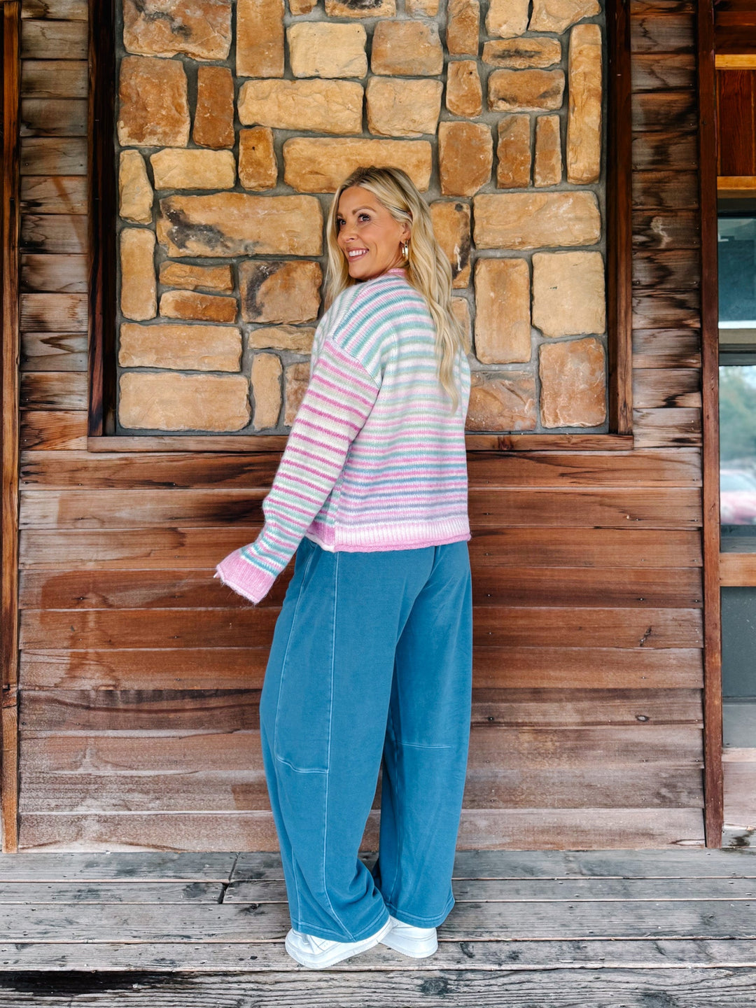 A woman stands on a rustic wooden porch in front of a stone wall, wearing a pastel striped knit sweater and wide-leg blue jeans.