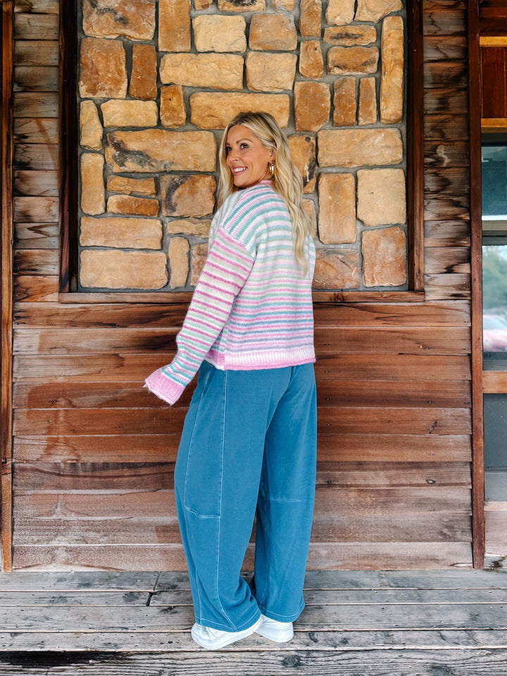 A woman stands on a rustic wooden porch in front of a stone wall, wearing a pastel striped knit sweater and wide-leg blue jeans.