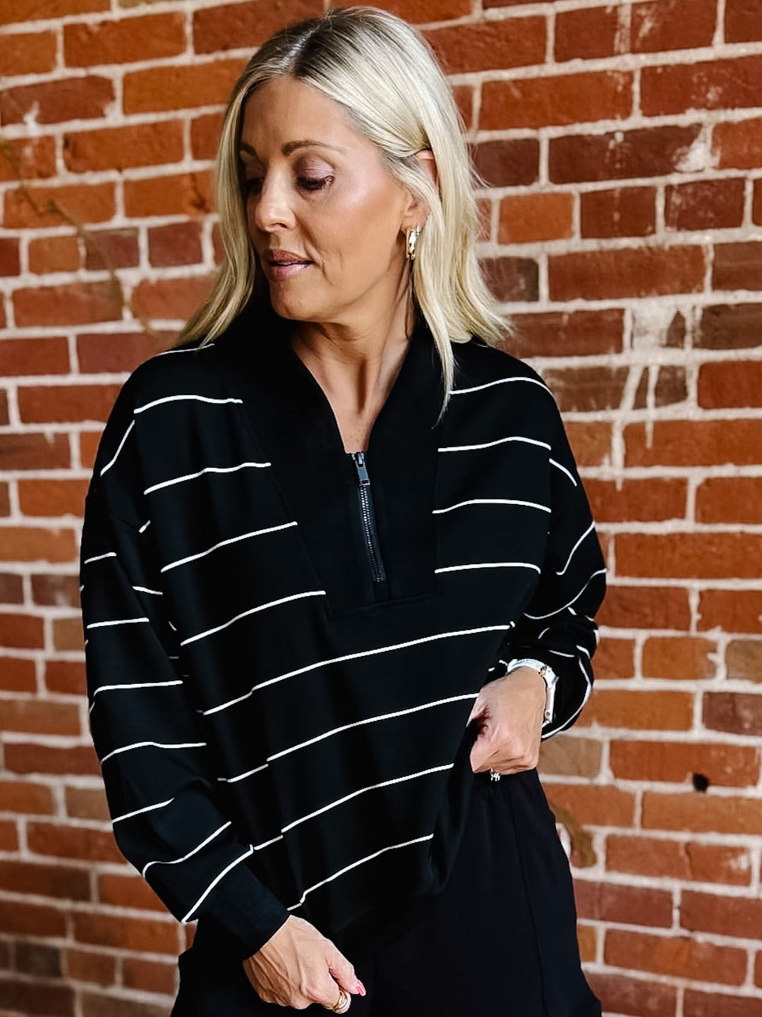 A woman with blonde hair wearing a black long-sleeve quarter-zip pullover with thin white horizontal stripes, standing in front of a red brick wall.