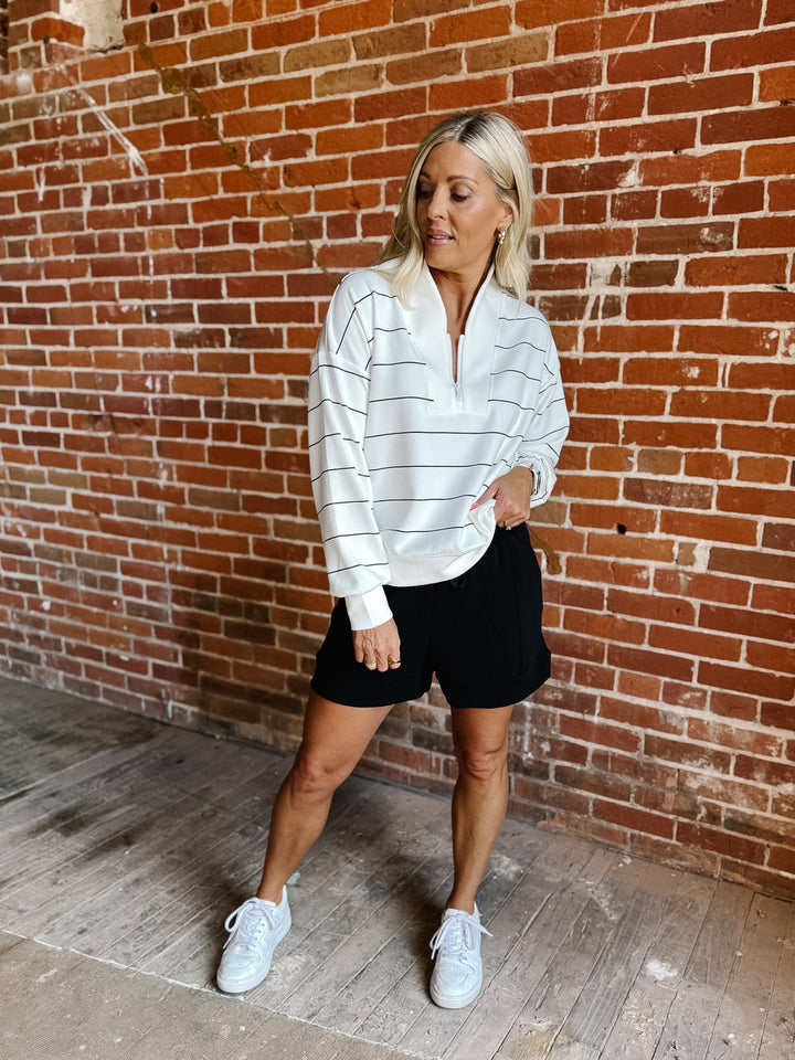 A woman posing in front of a red brick wall, wearing a white long-sleeve quarter-zip pullover with thin black horizontal stripes, black shorts, and white sneakers.