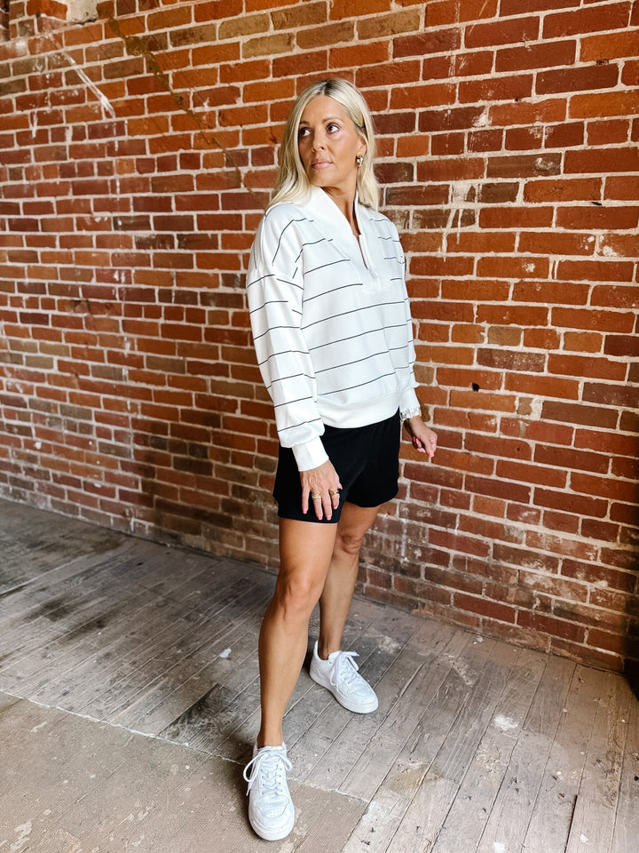 A woman stands in front of a red brick wall wearing a white long-sleeve collared shirt with thin dark horizontal stripes, black athletic shorts, and white sneakers.