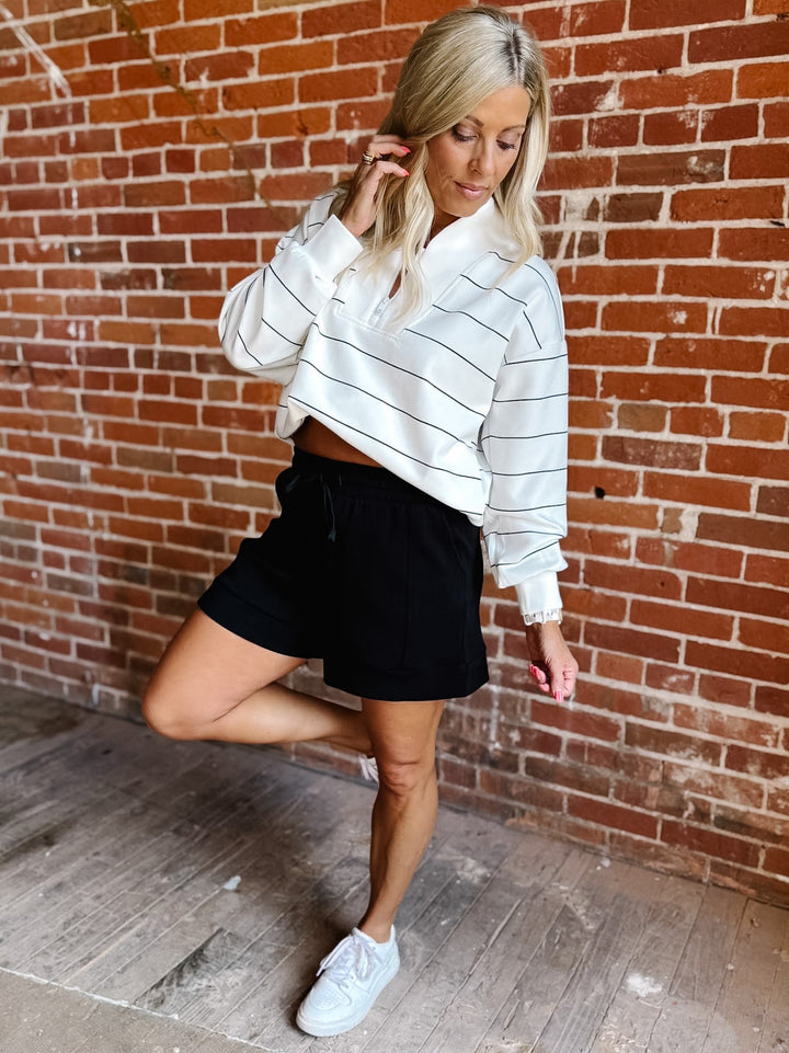 A woman posing in front of a red brick wall, wearing a white cropped sweatshirt with thin black horizontal stripes, black shorts, and white sneakers.