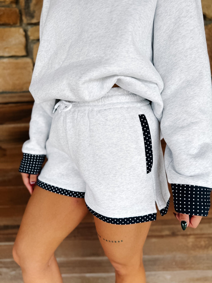 A person wearing light heather gray drawstring shorts with navy-and-white polka dot trim on the side pocket and hem, paired with a matching oversized sweatshirt. The photo is cropped from the chest down, showing the shorts and part of the sweatshirt in a real indoor setting with wood flooring and a stone wall.