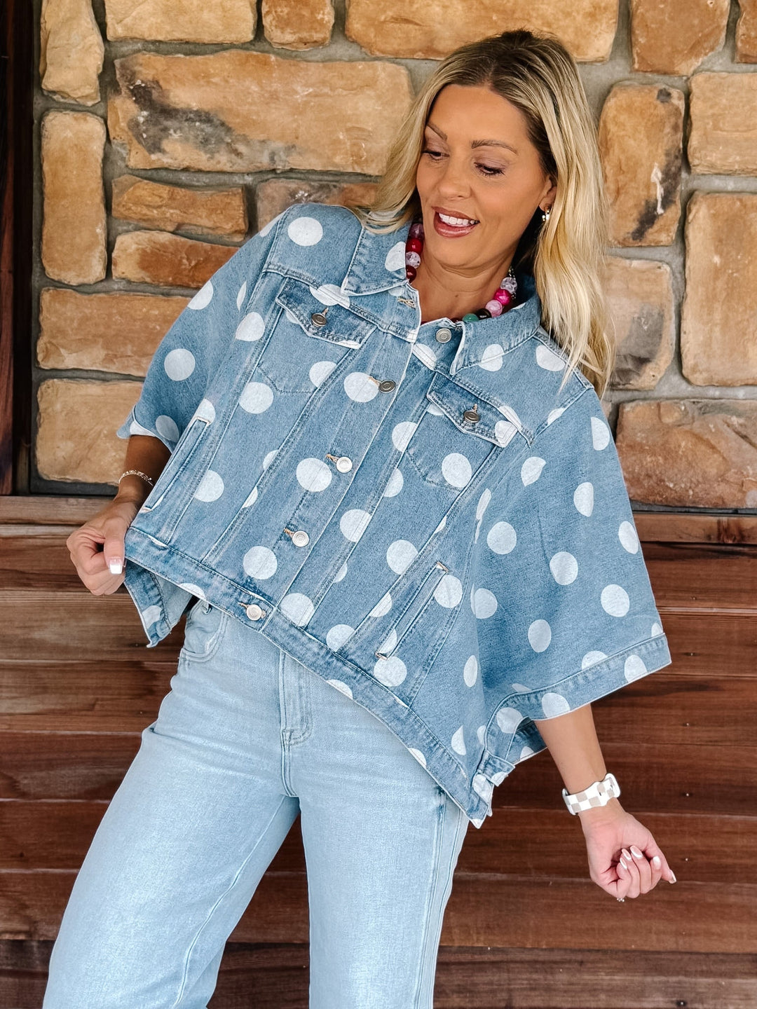 A woman wearing a light blue denim poncho-style jacket with white polka dots, paired with light-wash jeans, posing in front of a rustic stone and wood wall.