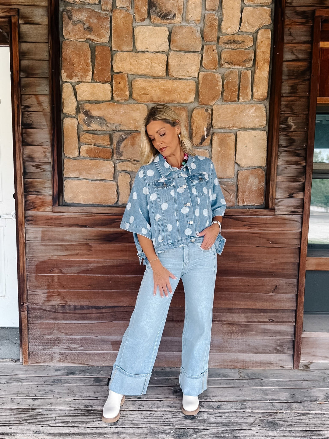 A woman stands on a rustic wooden porch in front of a stone wall, wearing a light blue denim jacket with white polka dots and matching light blue wide-leg jeans.