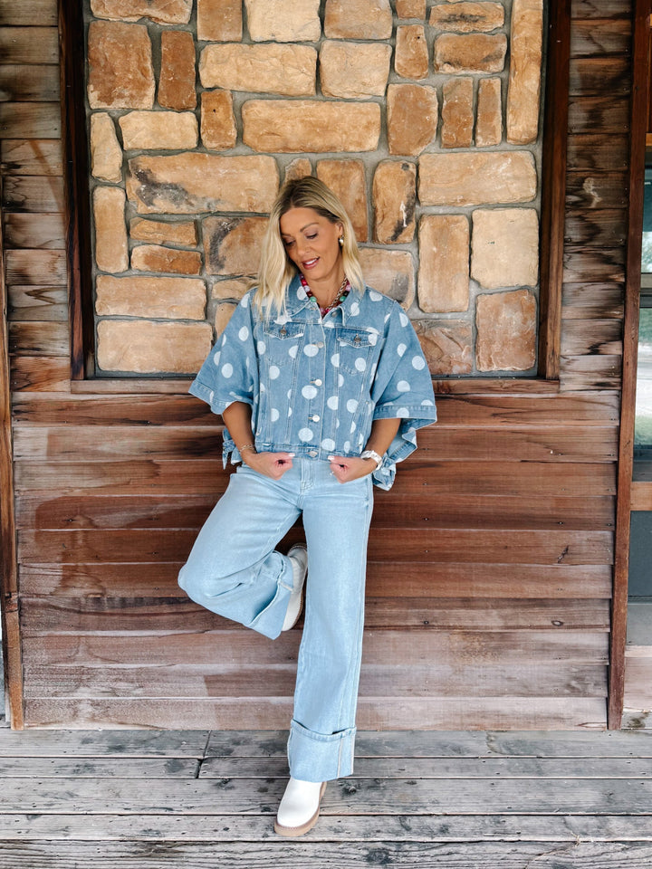 A woman posing in a light blue denim shirt with white polka dots and matching wide-leg light blue jeans, standing against a rustic stone and wood wall.