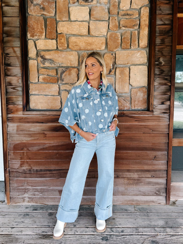 A woman stands on a wooden porch in front of a stone wall, wearing a light blue denim shirt with white polka dots and light blue wide-leg jeans.