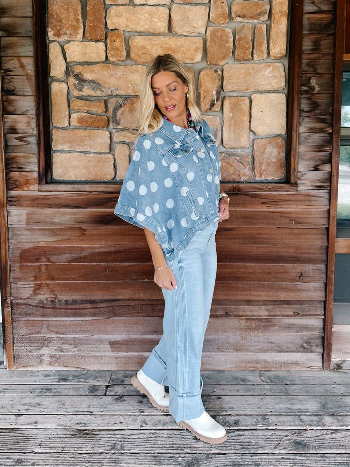 A woman stands on a rustic wooden porch in front of a stone wall, wearing a light blue denim poncho-style top with white polka dots, light-wash jeans, and white boots.