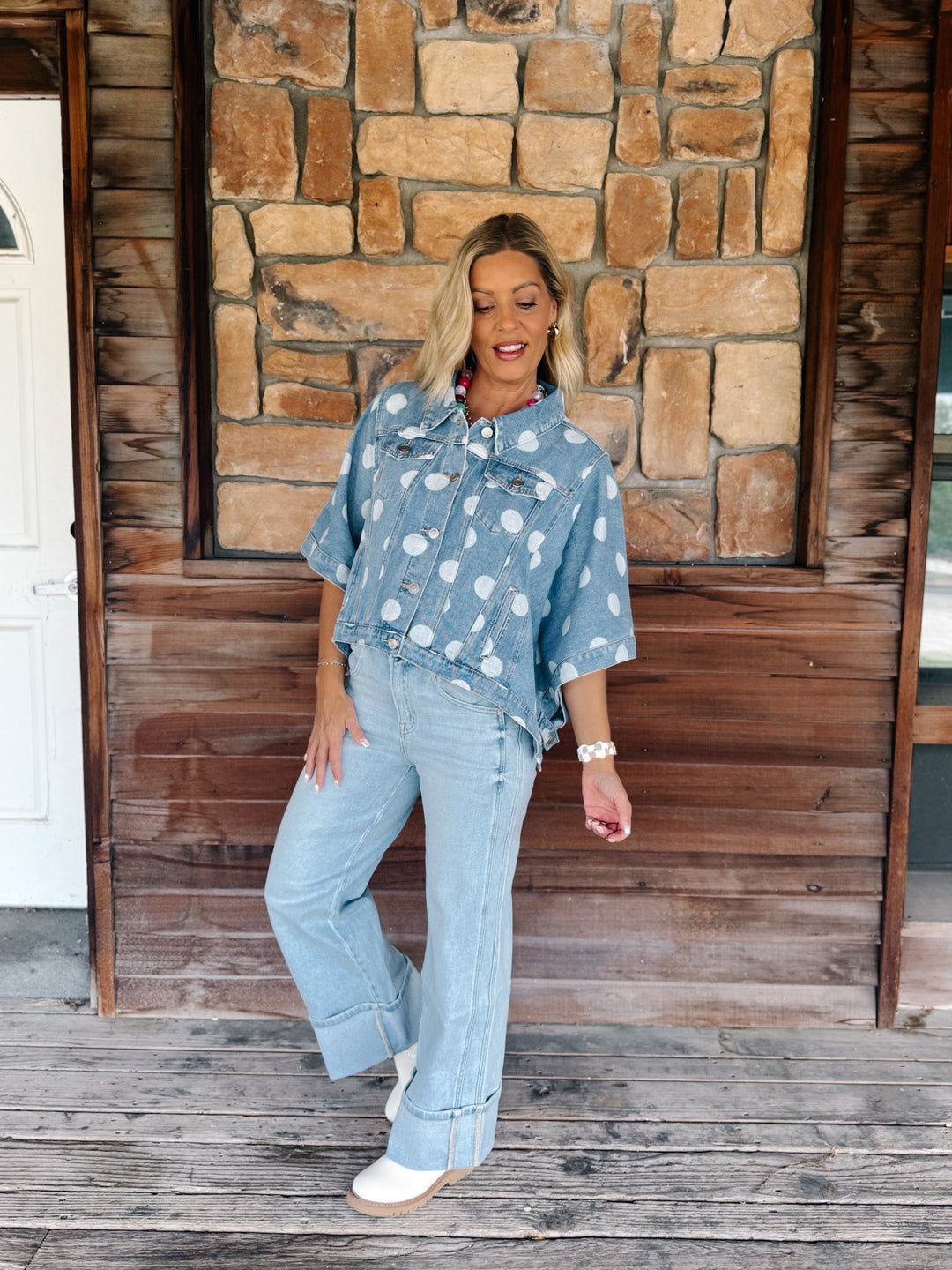 A woman stands on a wooden porch in front of a stone and wood wall, wearing a light blue denim shirt-jacket with a white floral print, light-wash flare jeans, and white shoes.