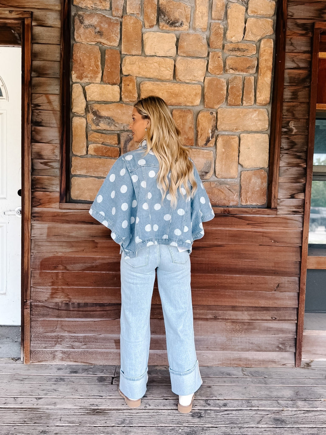 A woman stands on a rustic wooden porch in front of a stone wall, wearing a light blue denim jacket with white polka dots and light-wash wide-leg jeans.