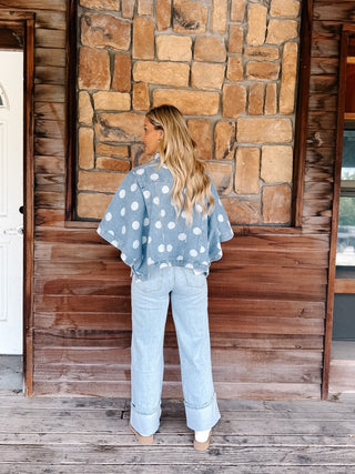 A woman stands on a rustic wooden porch in front of a stone wall, wearing a light blue denim jacket with white polka dots and light-wash wide-leg jeans.