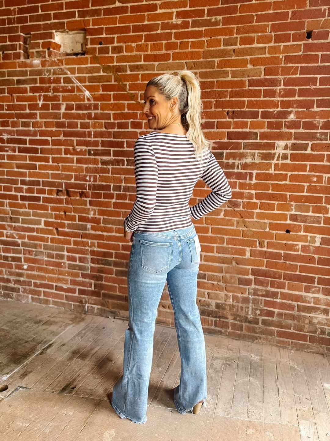 A woman wearing a brown and light blue horizontally striped long-sleeve boat-neck top, paired with blue jeans, standing against a red brick wall.