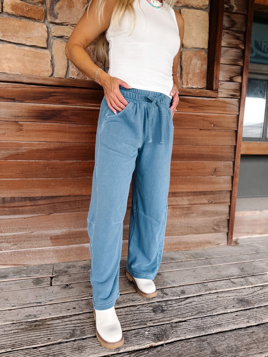 A person wearing light blue wide-leg drawstring pants, a white sleeveless top, and white boots, standing on a wooden deck in front of a rustic wood and stone wall.