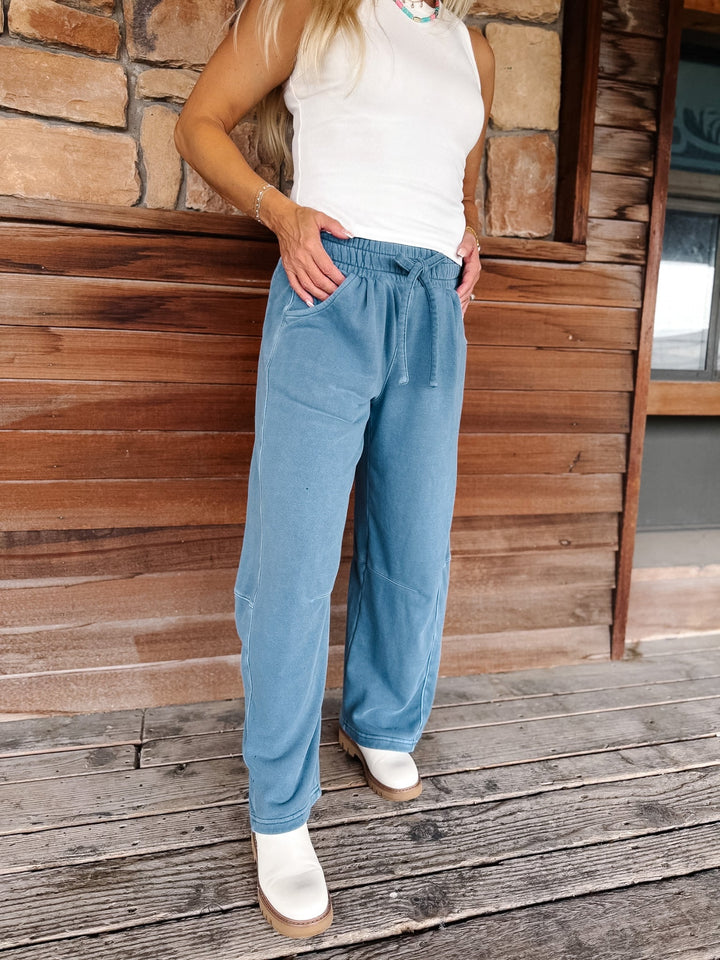 A person wearing light blue wide-leg drawstring pants, a white sleeveless top, and white boots, standing on a wooden deck in front of a rustic wood and stone wall.